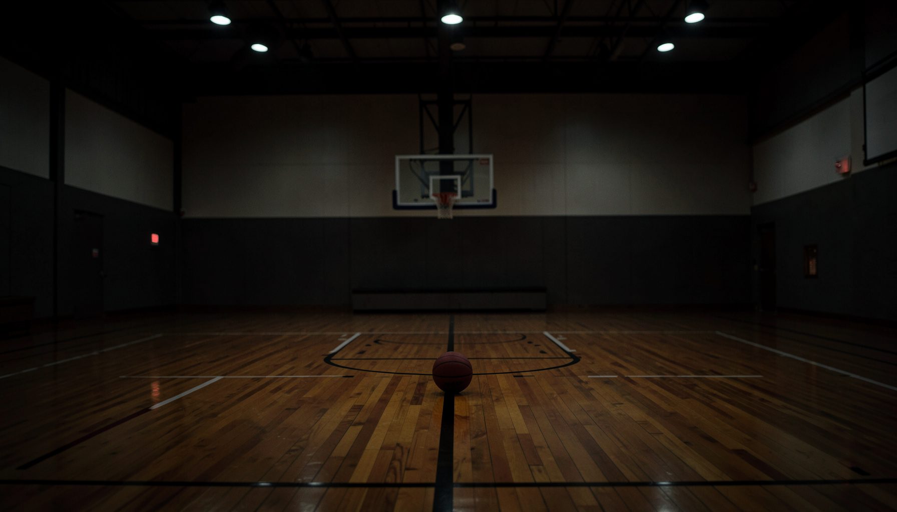 A dimly lit indoor basketball court with polished hardwood floors, a lone basketball resting near center court, and soft overhead lights casting a focused glow across the key.