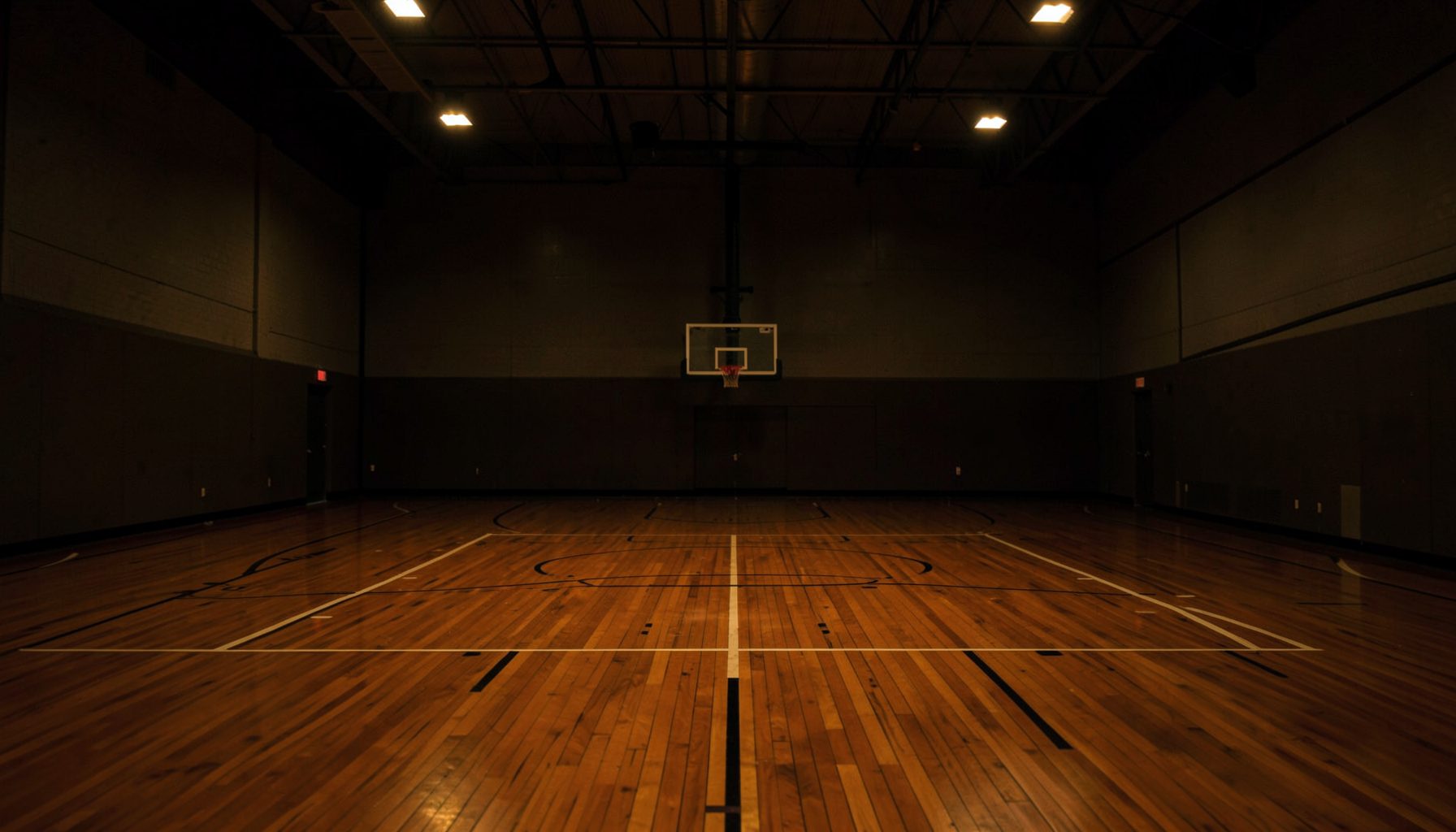 A dimly lit indoor basketball court with polished hardwood flooring, a backboard and rim in the distance, and warm overhead lights casting a focused glow on the playing surface.
