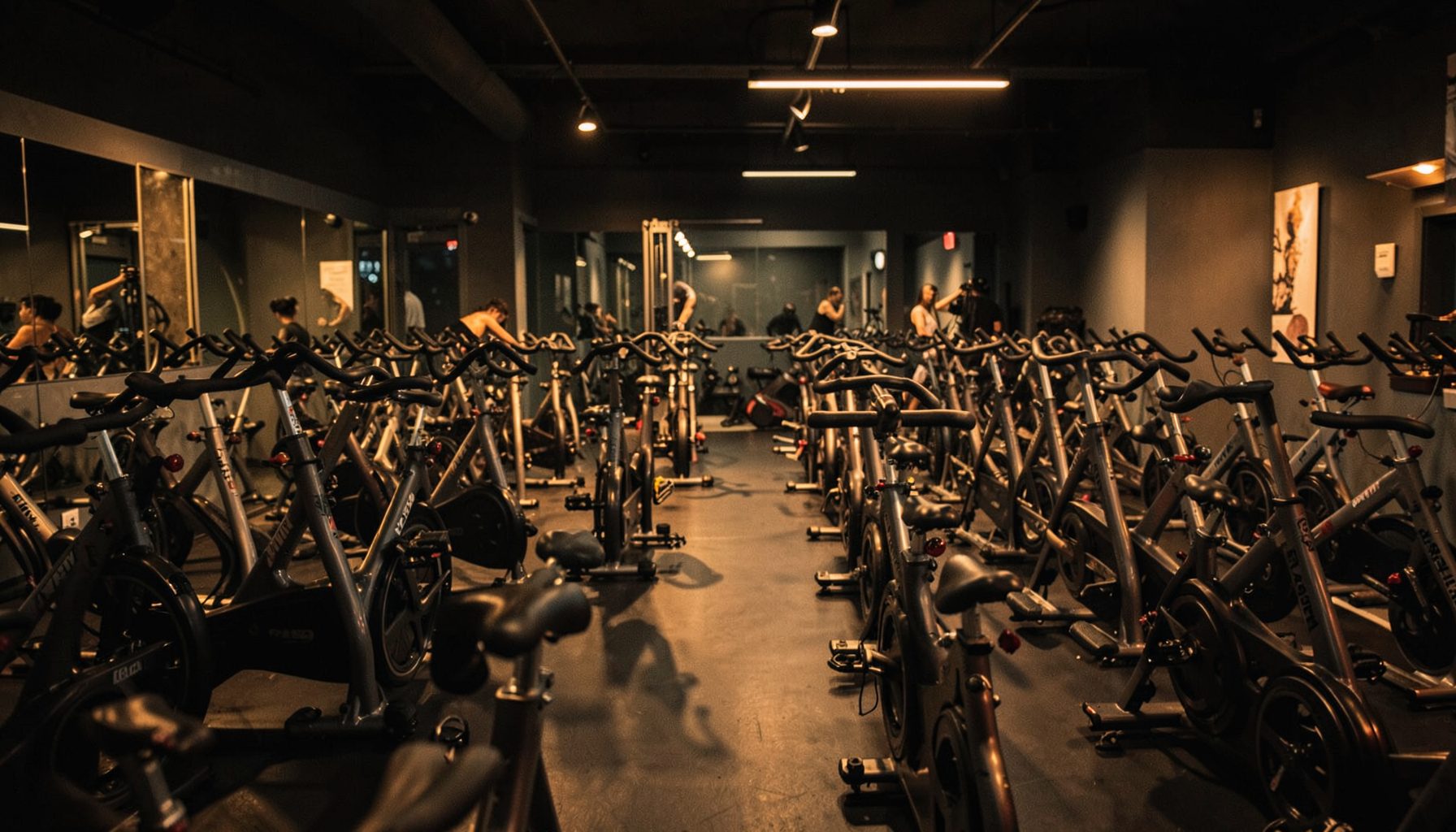 A dimly lit indoor cycling studio with rows of stationary bikes, warm amber and charcoal tones, energetic atmosphere