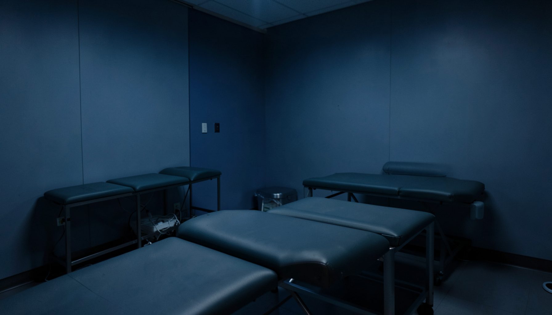 A dimly lit recovery room with soft blue and charcoal tones, featuring padded treatment tables and a calm, restorative atmosphere.