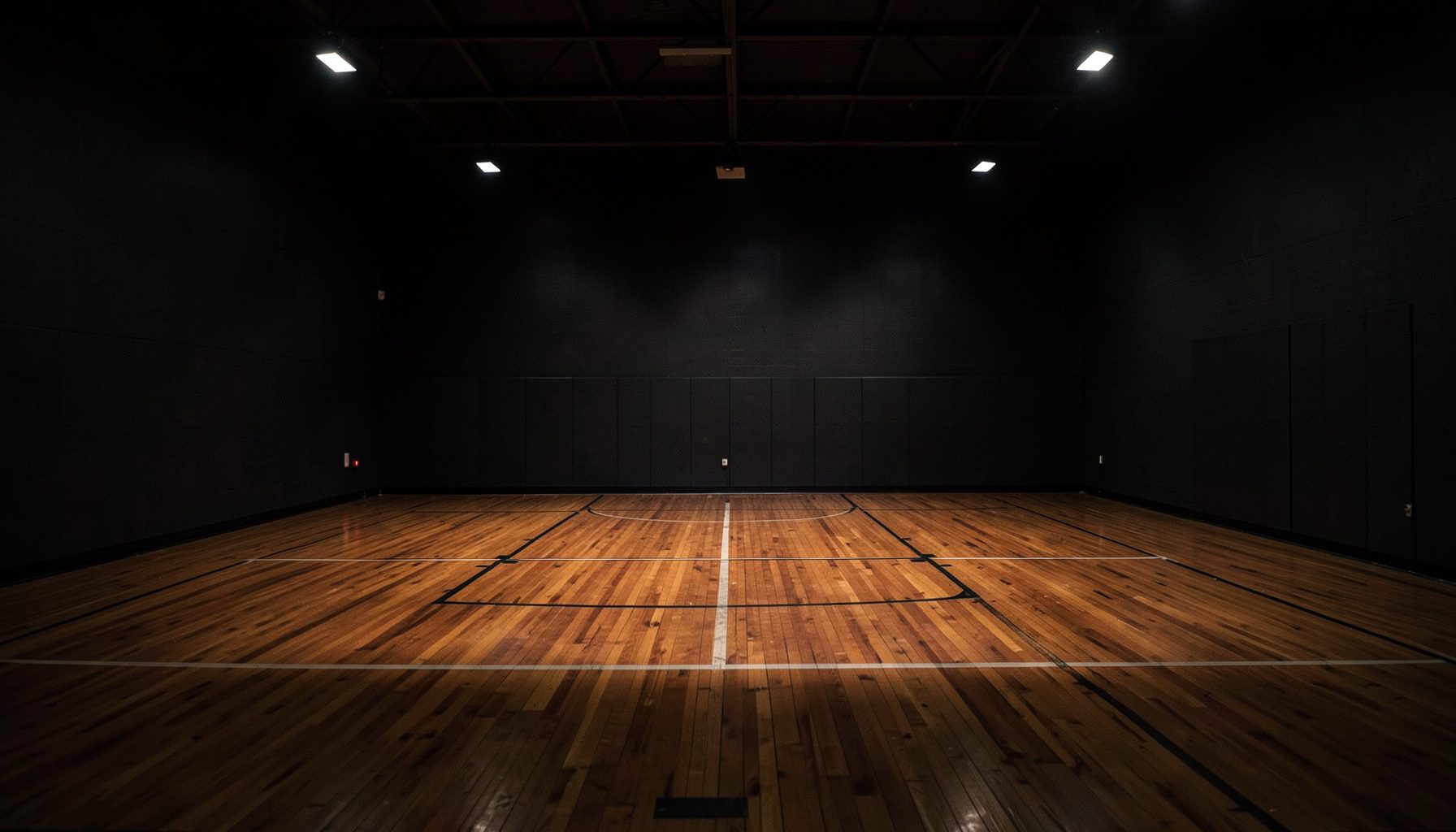 Dimly lit basketball court with polished hardwood floor, dark charcoal walls, and dramatic overhead lighting casting long shadows across the empty court