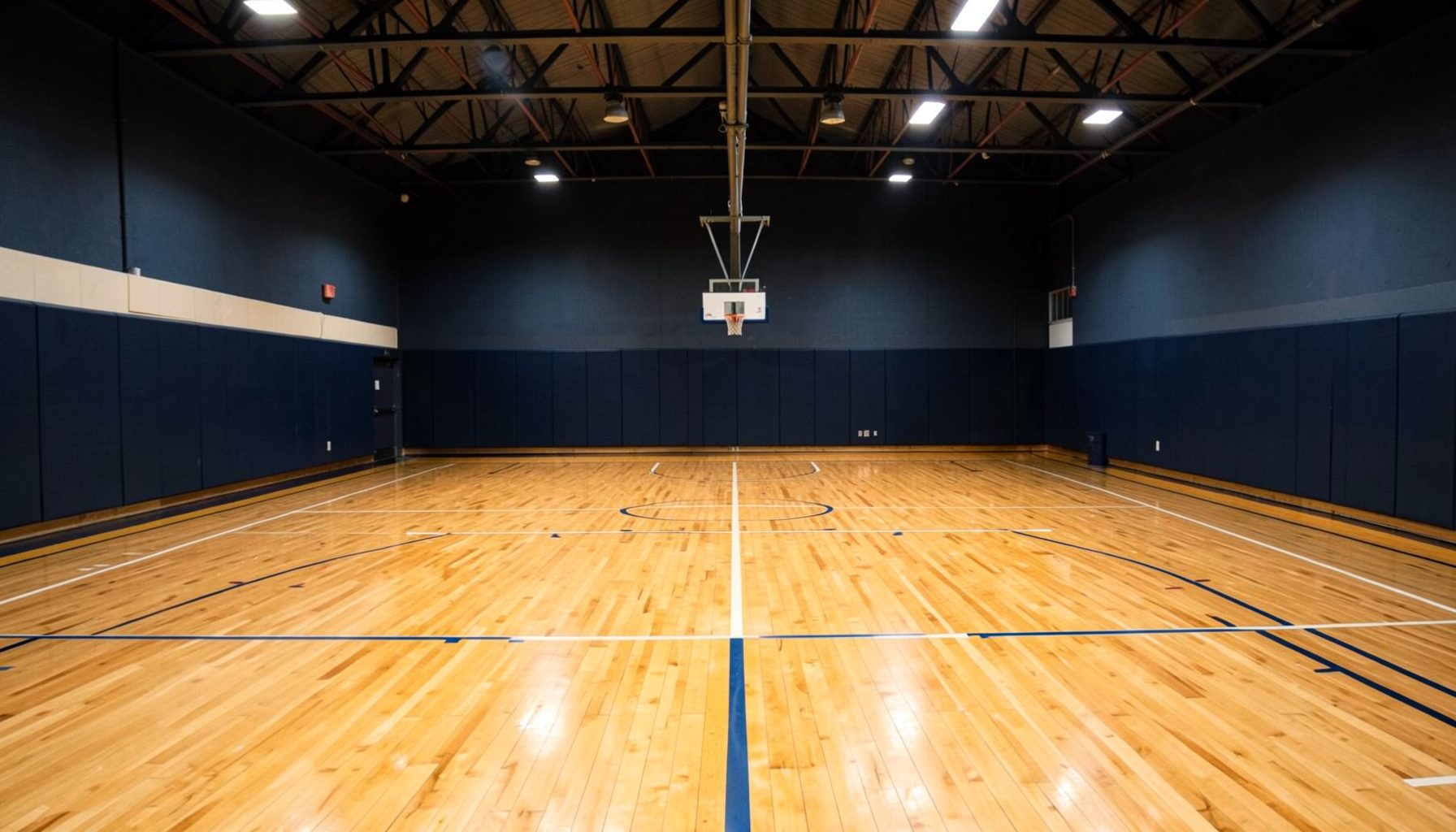 Indoor basketball court with warm wood flooring, dark navy walls, and overhead gym lighting, conveying an energetic athletic atmosphere