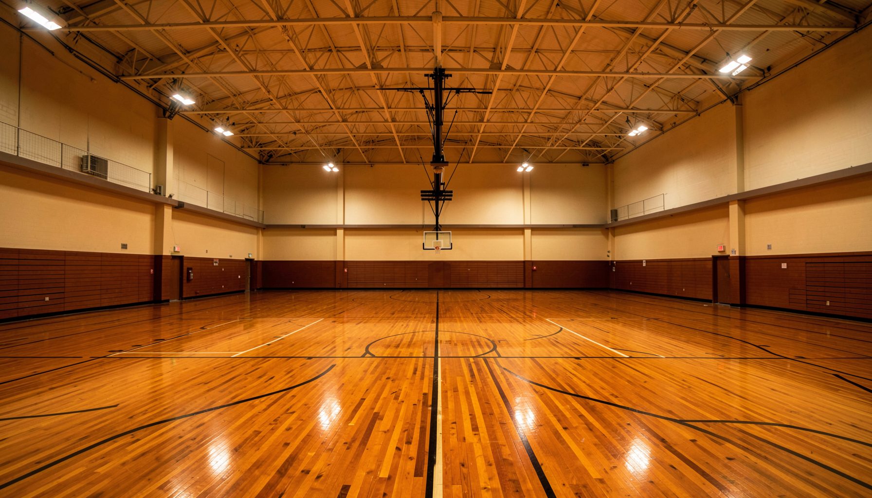 Wide-angle interior of a basketball gymnasium with polished hardwood floors, bright overhead lights, and empty courts stretching across the frame in warm amber and deep brown tones.