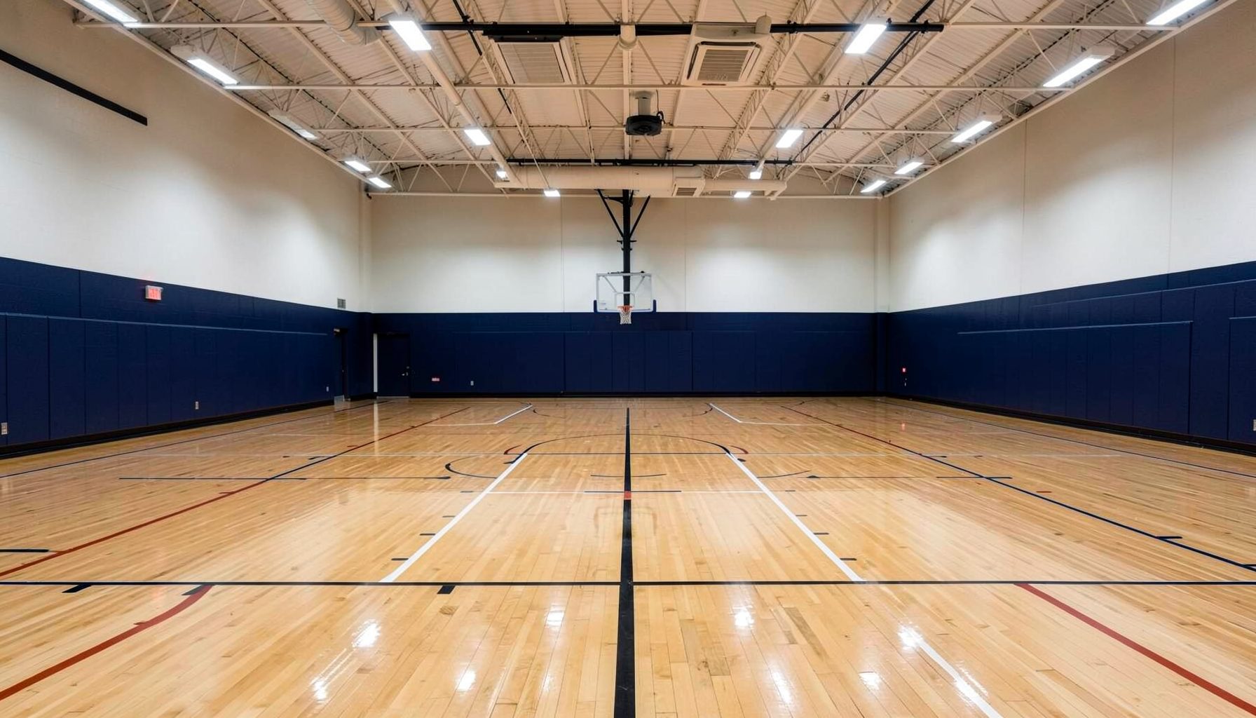 Wide interior of a modern athletic facility with polished hardwood basketball courts, bright overhead lights, and deep navy wall accents