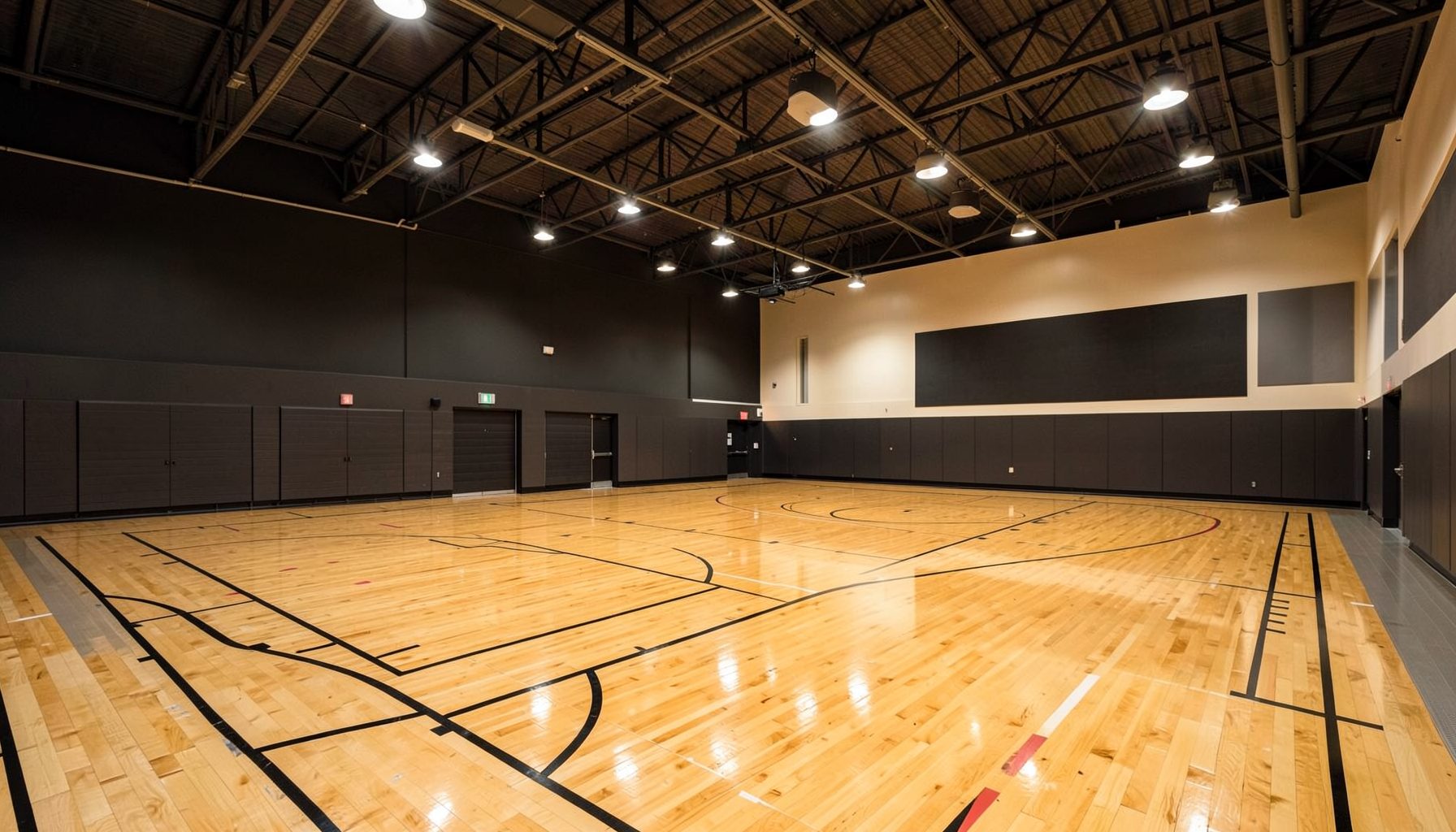 Wide interior view of a modern athletic facility with polished hardwood basketball courts, warm overhead lighting, and deep charcoal and cream tones throughout the open space.
